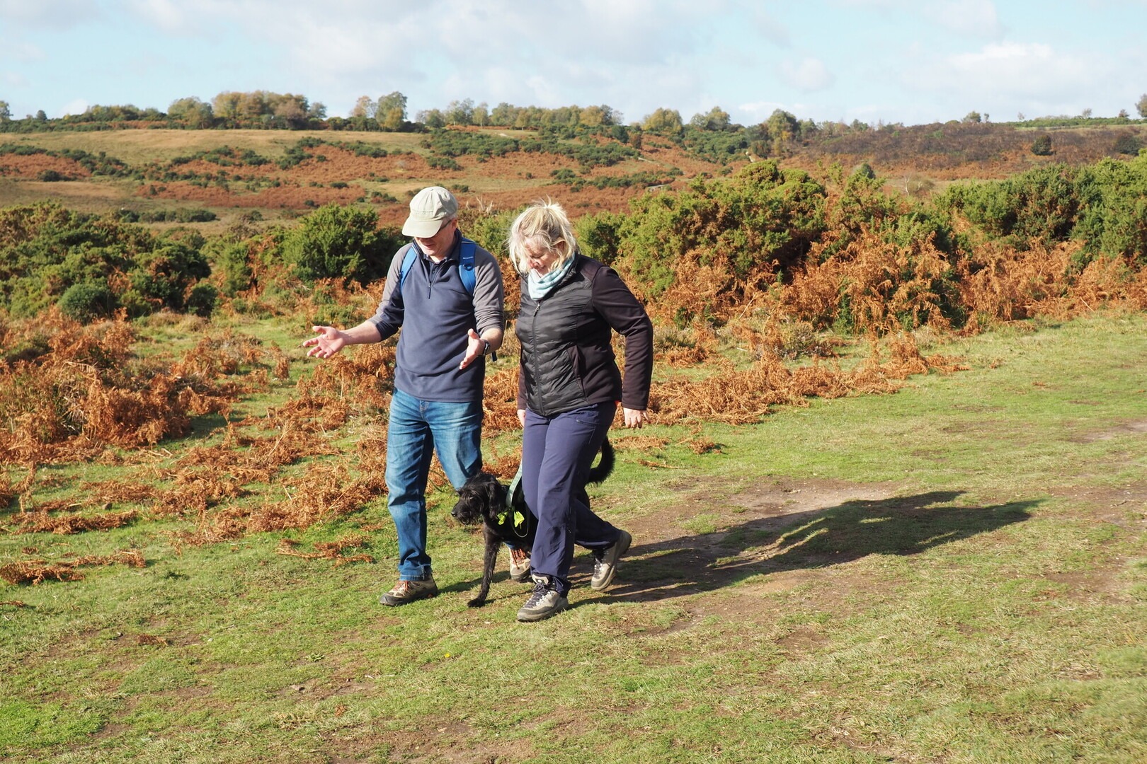 Two hikers walk a black dog along a grassy path in the New forest.