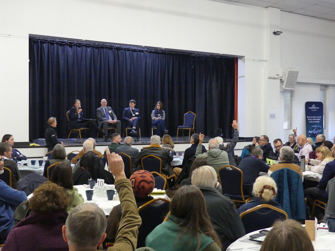 A panel discussion on a stage in a village hall. The room is full of people seated and some have their hands raised to ask a question.