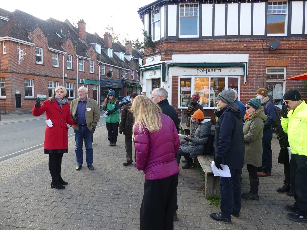 A group of people in a village centre one is addressing the group and pointing