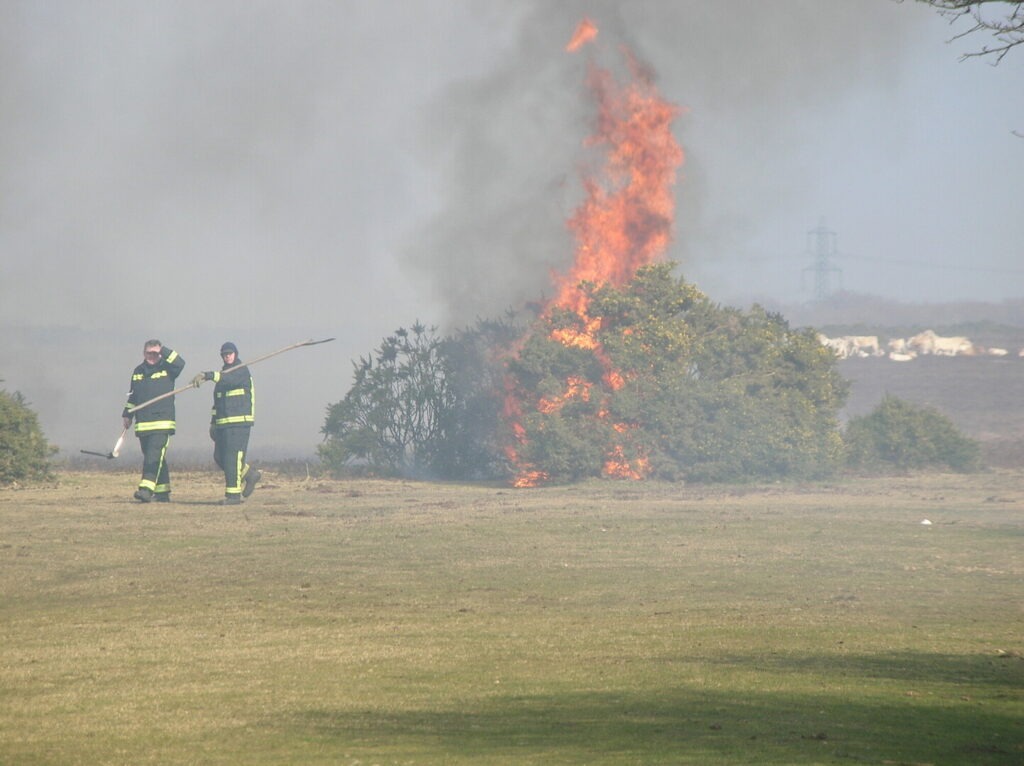 Two firefighters with tools walk across a field as a large bush fire burns behind them, sending flames and smoke into the air