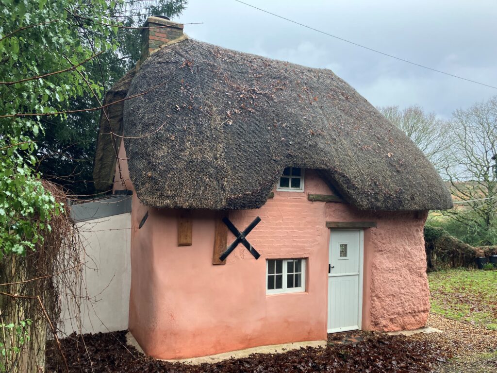 Pink thatched-roof cottage with white door and small windows, brick chimney, and wooded countryside surroundings