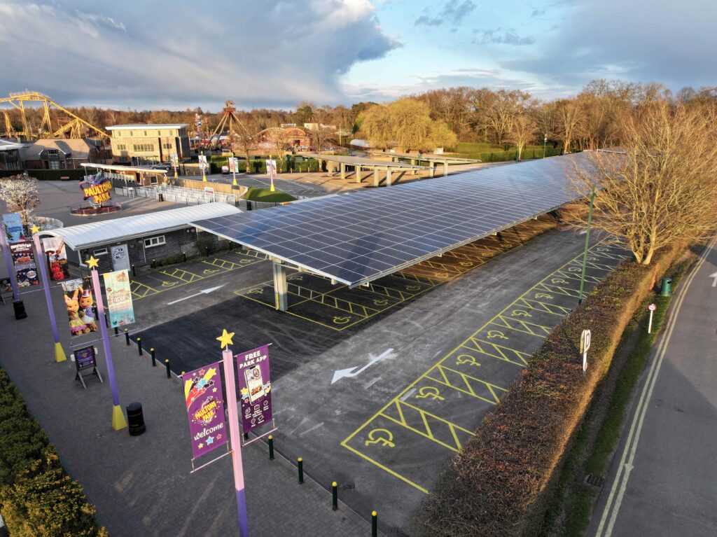 Solar panel carport canopy over marked parking bays at Paultons Park entrance, with park buildings and rides in the background