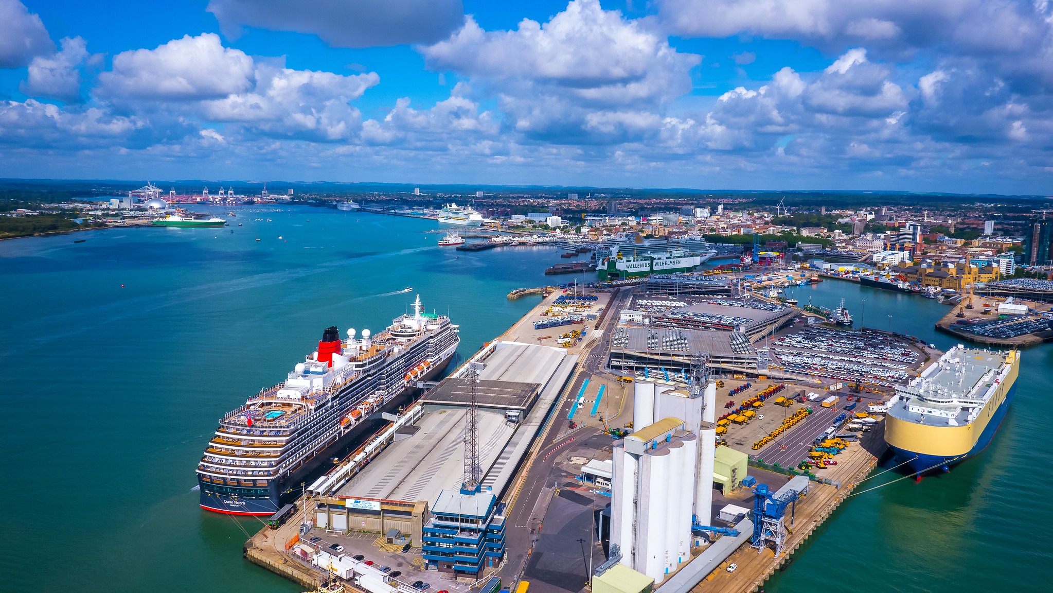 Aerial view of Southampton port with cruise ships docked beside cargo terminals and car storage yards under a blue sky with clouds