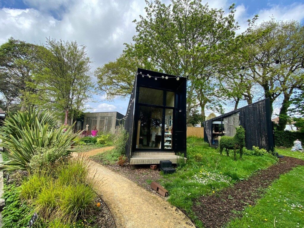 A garden space with a gravel path lading up to a small art studio. The studio is tall, has black outside walls and a large glass floor to ceiling window on the front side.