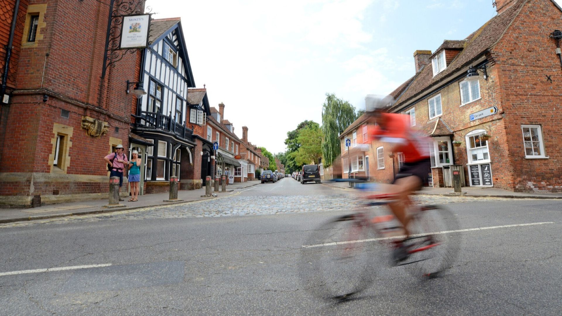 Blurred cyclist in red riding past brick and timber buildings on a village street in Beaulieu, with two pedestrians on the left sidewalk