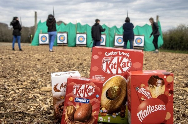 Easter chocolate boxes in foreground with archers aiming at targets on an outdoor range behind green netting