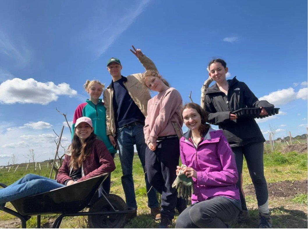 A group of young people on a farm on a sunny day. One is kneeling down, one is sat in a wheelbarrow and the rest are standing.