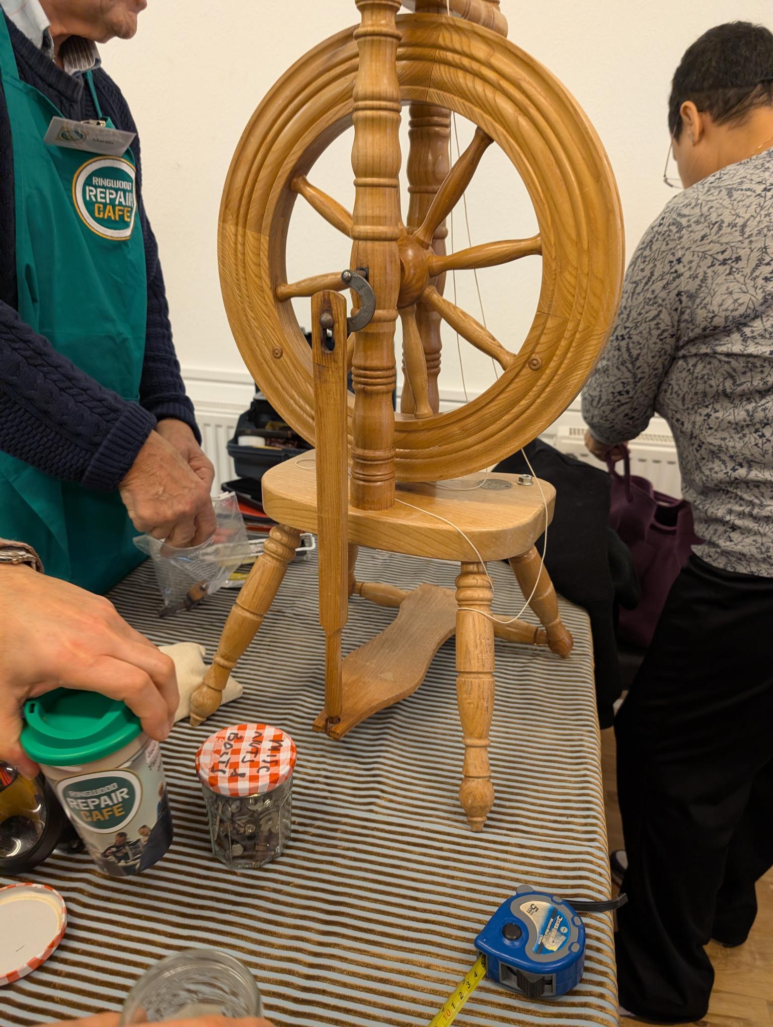 Old fashioned wooden spinning wheel in the centre of a table, being repaired and inspected by 2 people standing either side