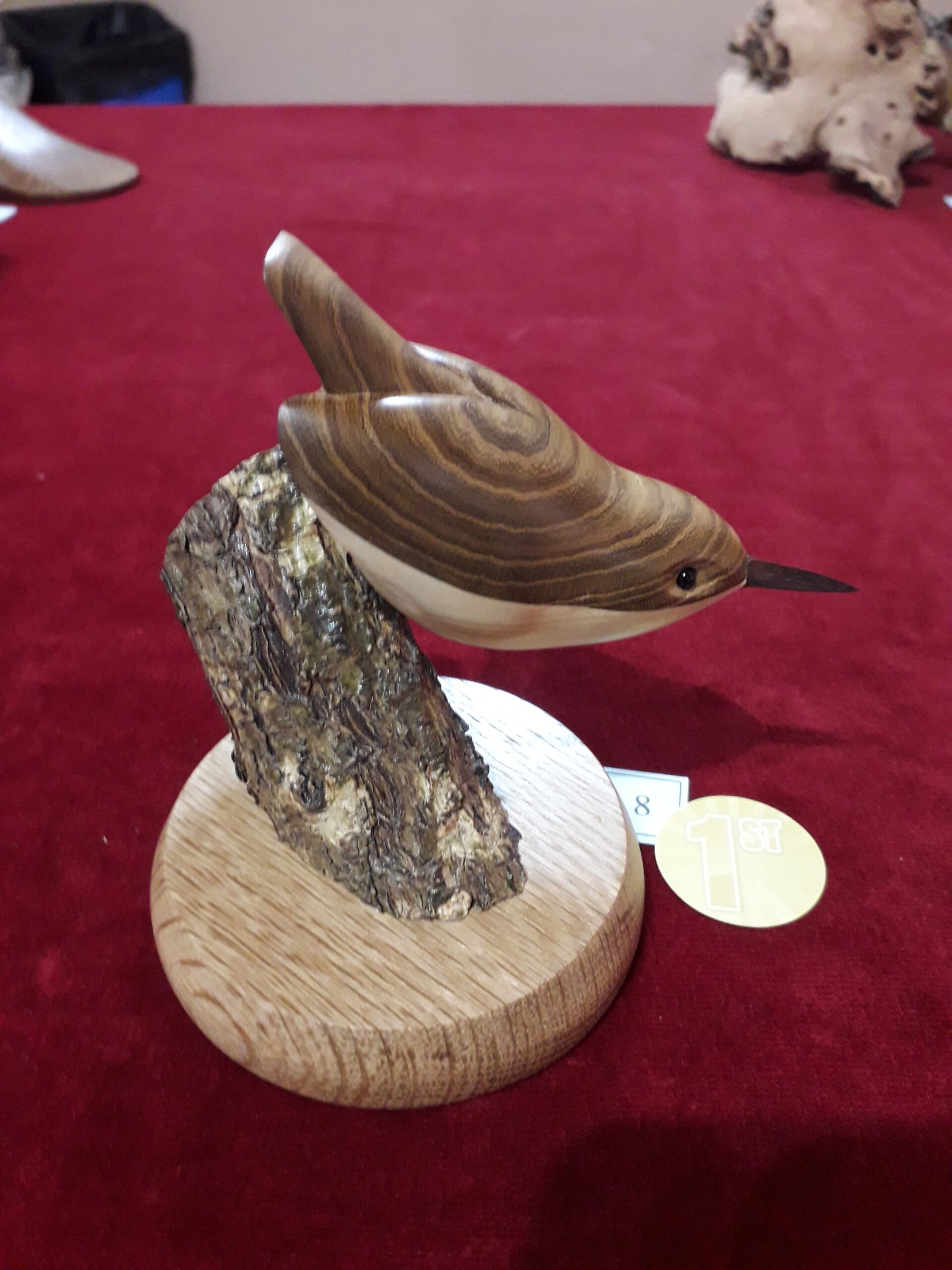 Carved wooden bird sculpture perched on a bark-covered branch base on a red table, with a gold 1st place badge nearby