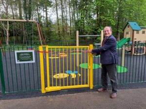 Man cuts a red ribbon at the yellow gate of the Queen Elizabeth II play area, with swings and a slide behind him