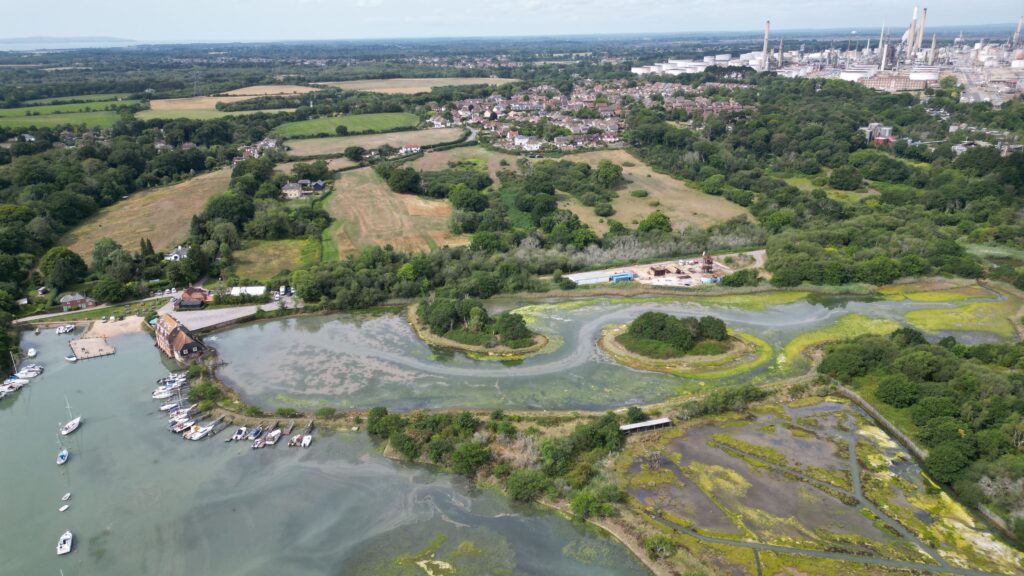 Aerial view of tidal creek and marsh with boats by a quay, woodland and fields, village housing, and an industrial refinery in distance