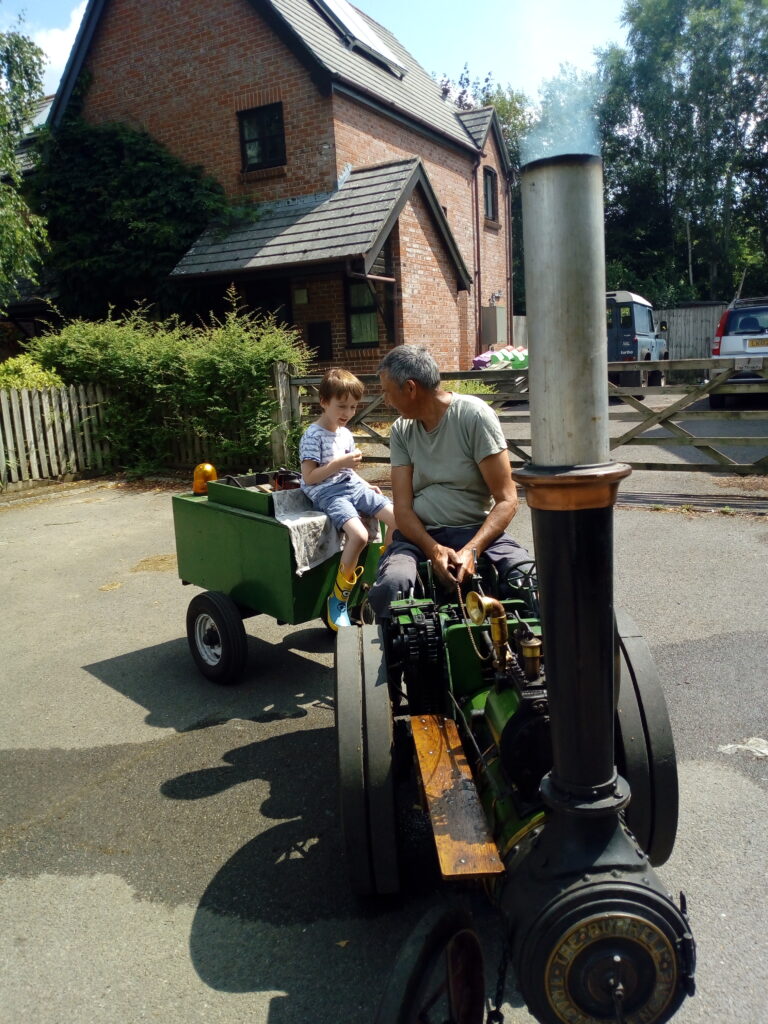 Man and child sitting on a small steam traction engine with green cart, smoke rising, in front of a brick house and gate