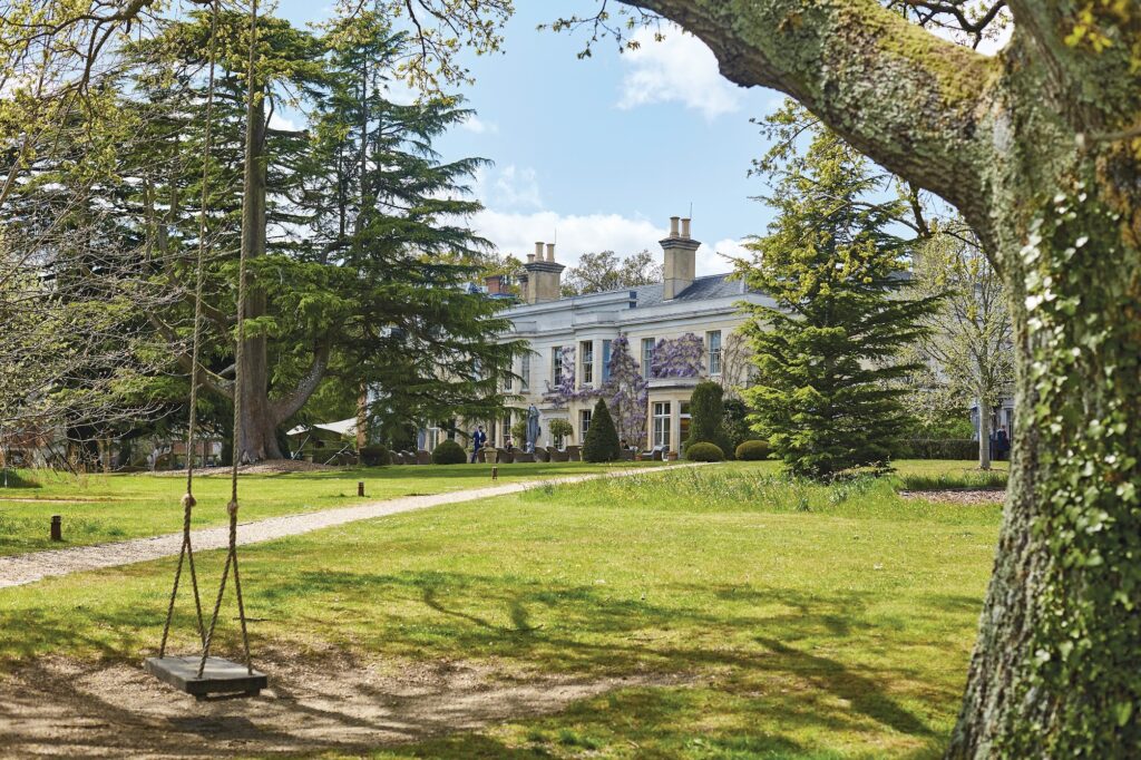 Country house hotel with wisteria on the facade, viewed across a lawn with a gravel path, tall trees, and a wooden swing in foreground
