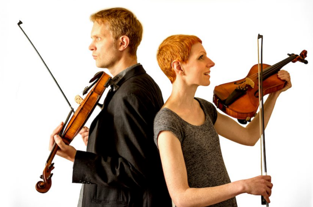 Two violinists standing back to back, holding violins and bows against a white background