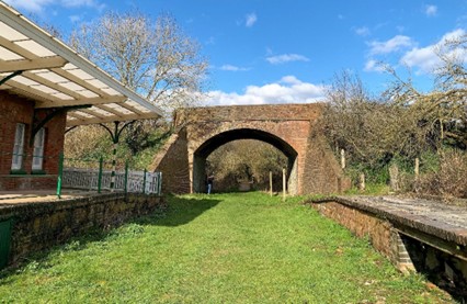 Disused brick railway bridge over grass-covered track bed, with old station platform and canopy on the left under blue sky