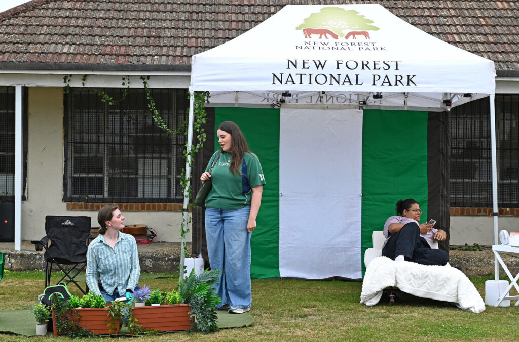 Three actors take part in a performance of Bringing the Outside in during the 2025 New Forest Thrive festival. All three are in a field - one is on a bed, one kneeling behind a plant pot, and one standing.