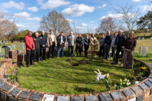 Group of people standing in a cemetery around a freshly dug hole for planting a sapling, with spades on the grass