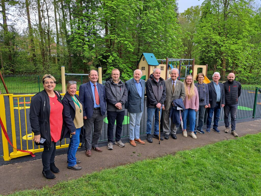 Group of 11 people posing by the fence at the Queen Elizabeth II play area opening in Ashurst, with playground equipment behind