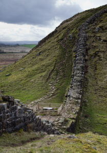Hadrian’s Wall running along a grassy hillside, with stone steps climbing the slope under a cloudy sky