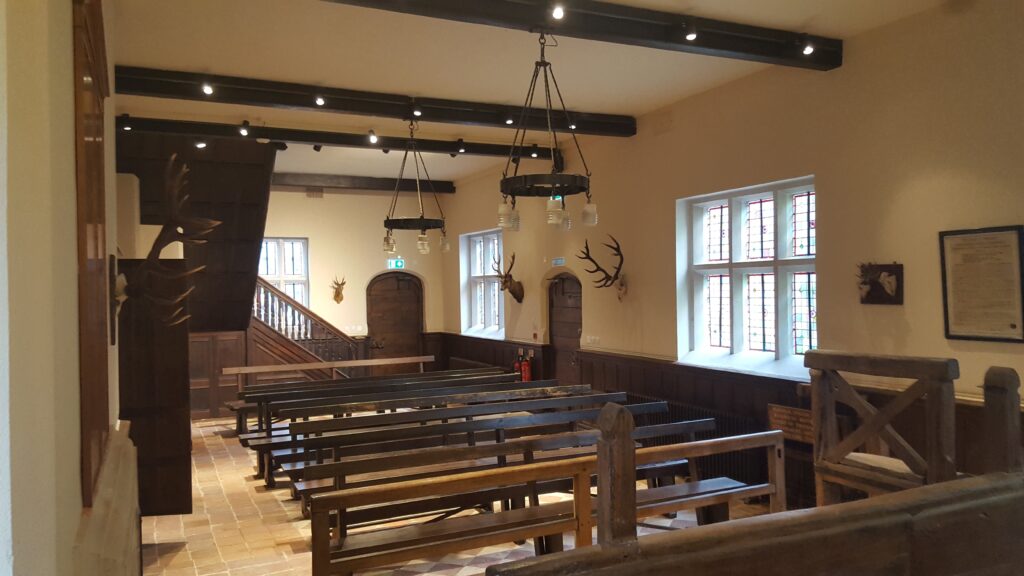 Interior of Verderers Court, Lyndhurst with wooden benches, dark ceiling beams, hanging lights, stained-glass windows, and mounted antlers