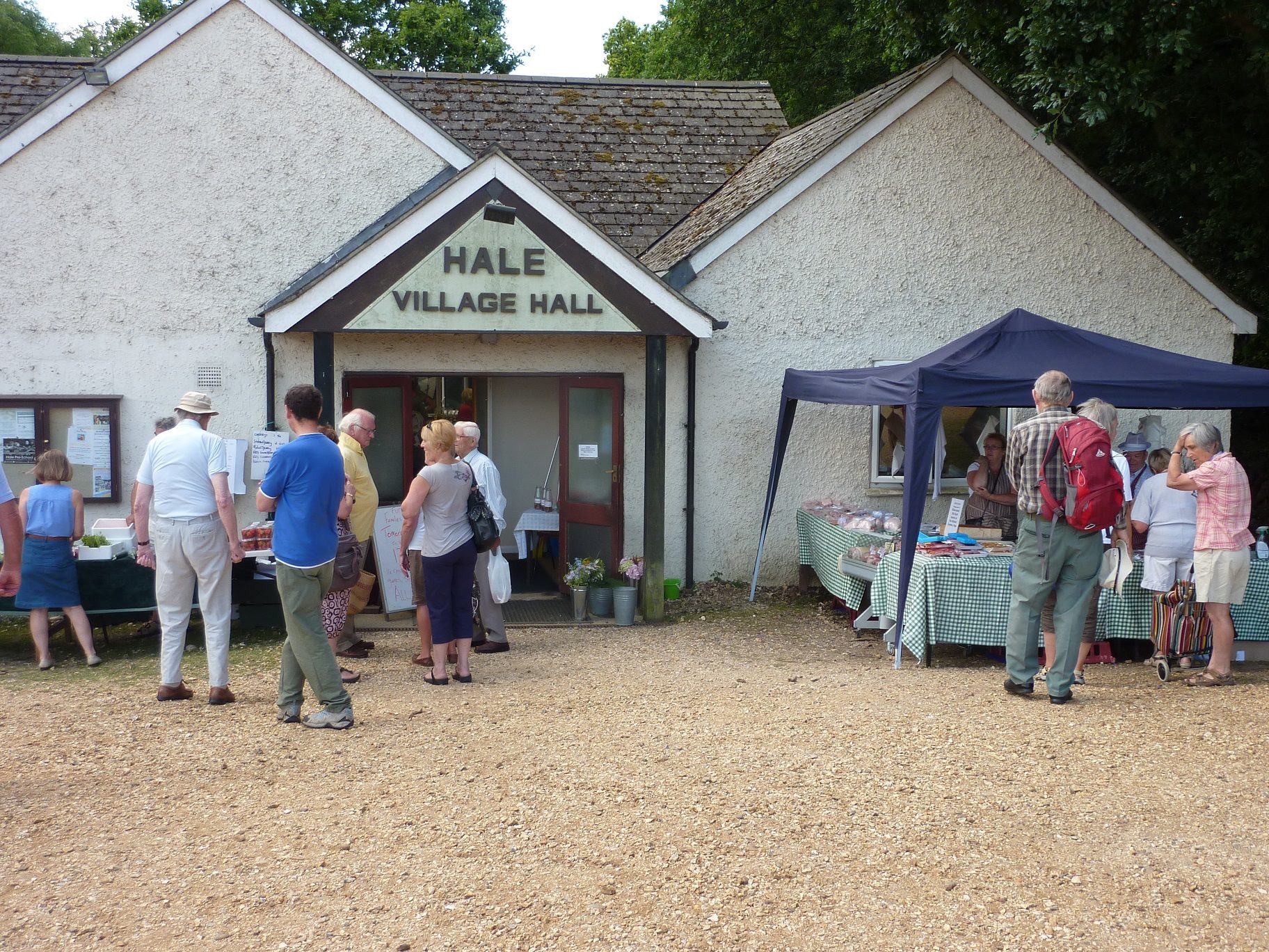 People browsing outdoor stalls outside Hale Village Hall, with a blue canopy over a table of goods