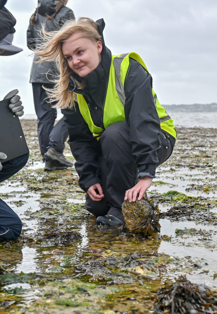A woman in high vis and waterproofs knelt on a beach at low tide on an overcast and windy day. She is lifting a rock to show young people the sea snails attached