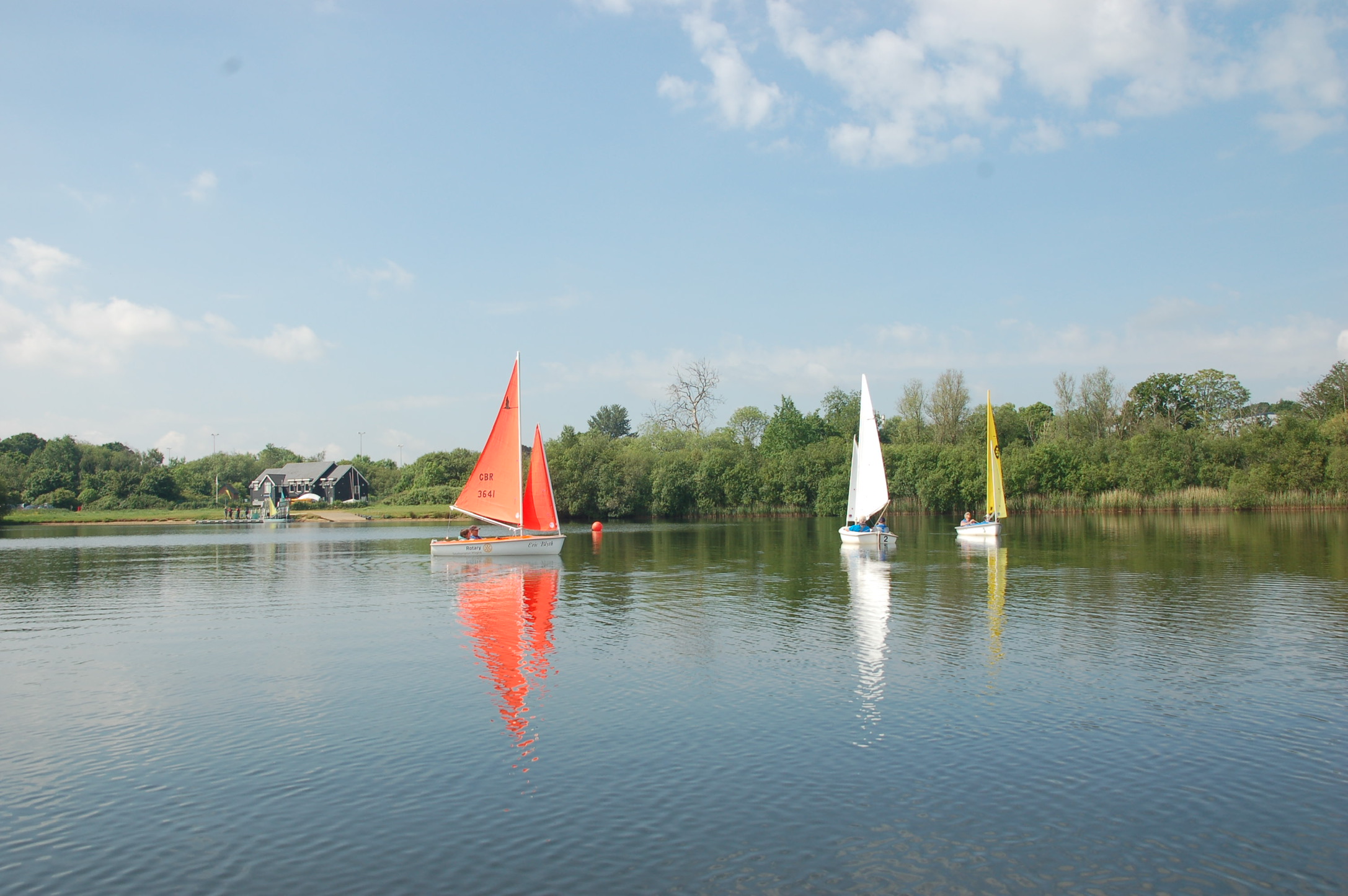 Three small sailboats with red, white, and yellow sails on a calm lake, reflecting in the water near a tree-lined shore