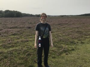 Young woman wearing a dark outfit, smiling and standing on grass with heathland and trees behind her.