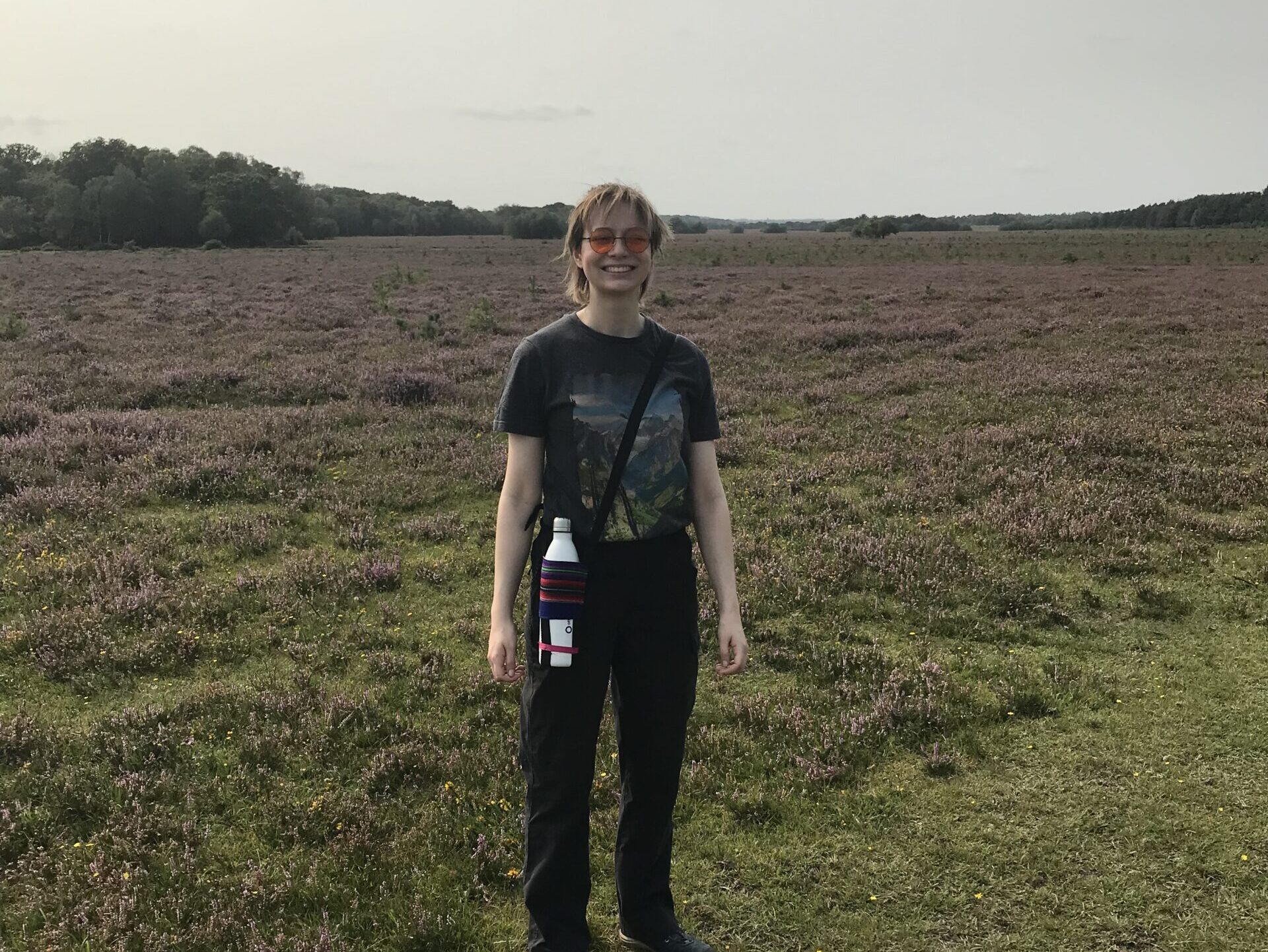 Young woman wearing a dark outfit, smiling and standing on grass with heathland and trees behind her.