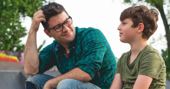 Man in glasses and green plaid shirt sitting outdoors, looking at a boy in a green T-shirt