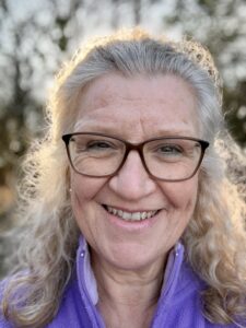 Close-up portrait of Fran Carpenter smiling, wearing glasses and a purple fleece outdoors with a blurred tree background