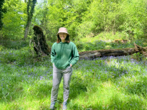Young woman stands smiling in a woodland area surrounded by trees and bluebells.