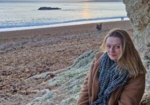 A young woman sitting on rocks to the right hand side of the image. In the background is a sandy beach, sea and dramatic sunset behind the clouds.