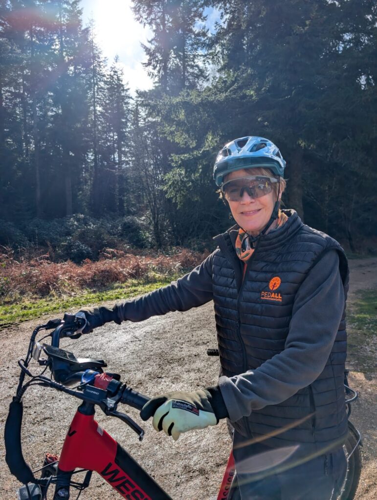 Christine Cooke, PEDALL – New Forest Inclusive Cycling Volunteer Ride Leader and a Ride Leader for PEDALL ladies 100-mile New Forest Loop challenge sits on a bike in a black jacket and orange PEDALL tshirt.