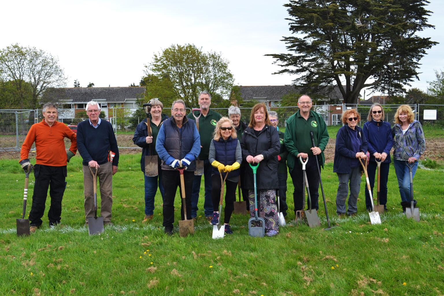 Group of 12 volunteers holding spades and tools on a grassy field, standing in front of a fenced garden area