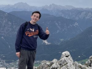 A young man stands on a rocky mountain top, with mountains in the background. He smiles and gives a thumbs up.