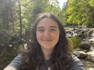 Smiling woman with long dark hair taking a selfie by a rocky stream in a sunlit green forest