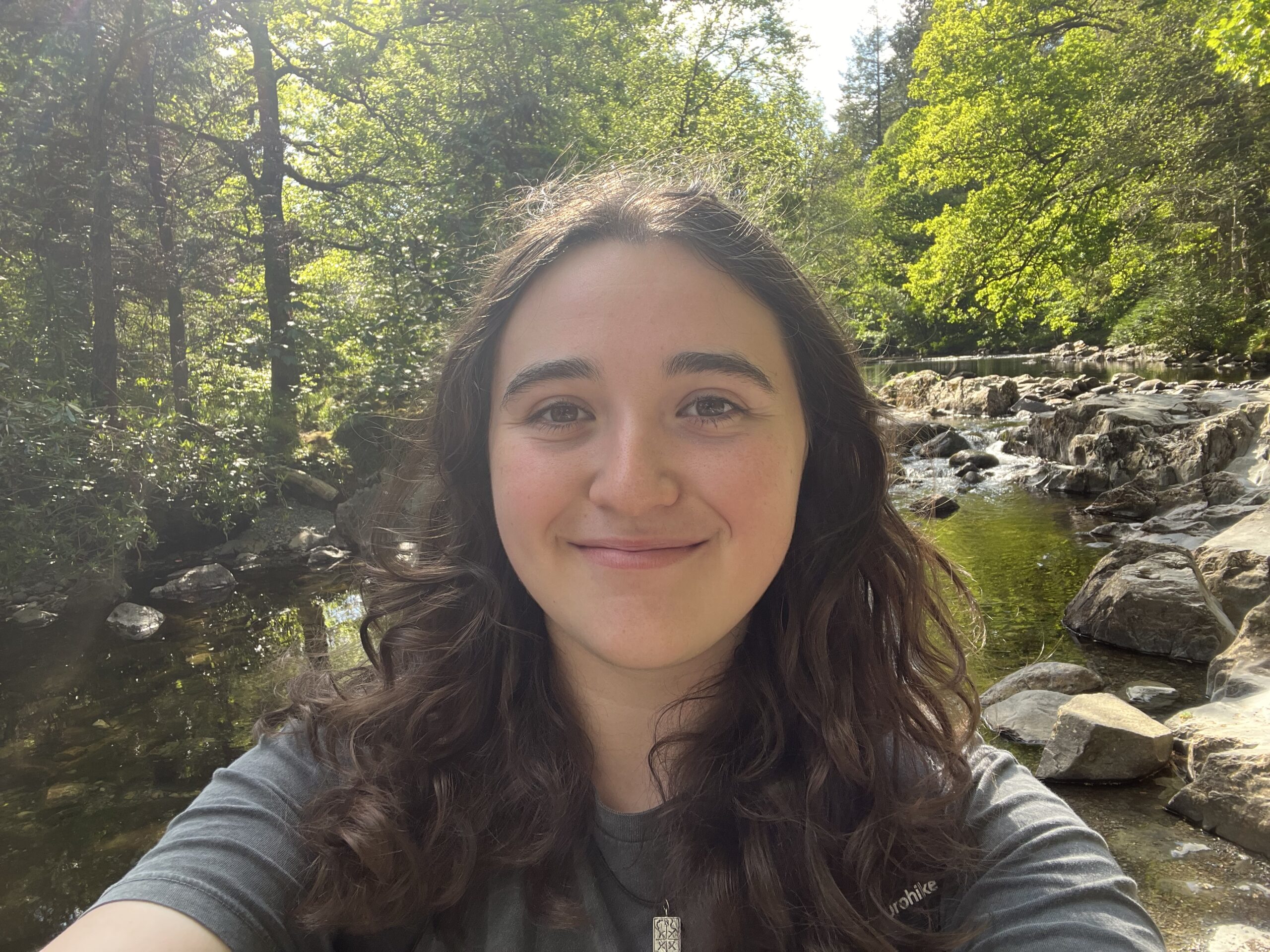 Smiling woman with long dark hair taking a selfie by a rocky stream in a sunlit green forest