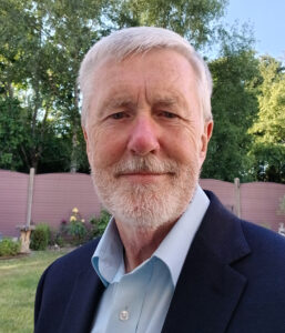 Close-up portrait of Martin Eyre, a gray-haired man with a short beard in a blazer and blue shirt, outdoors with trees behind