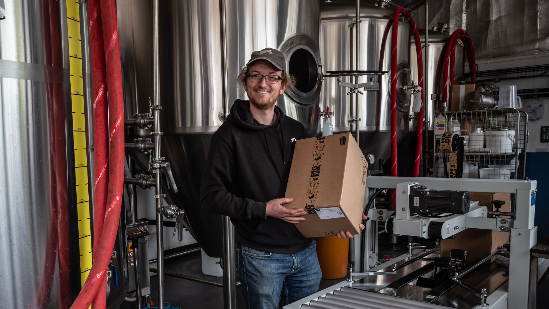 Worker holding a Pig Beer cardboard box inside a brewery, with stainless steel tanks, hoses, and packaging machinery behind him