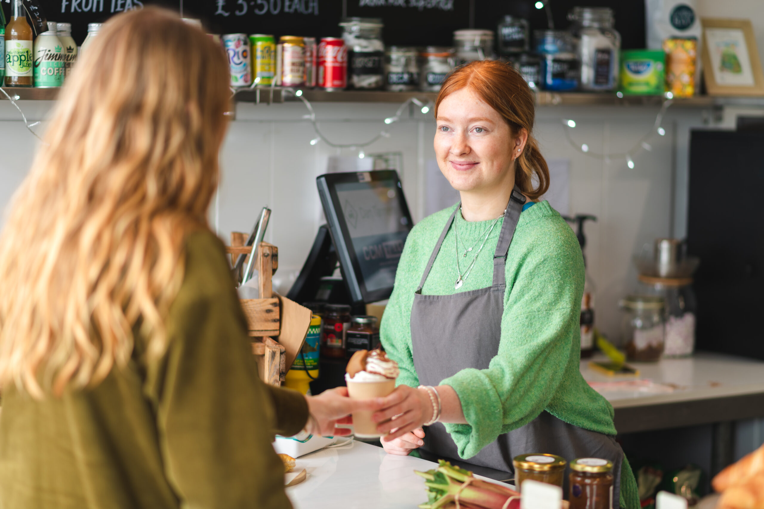 Cafe worker in a green sweater and apron hands a takeaway coffee to a customer at the counter