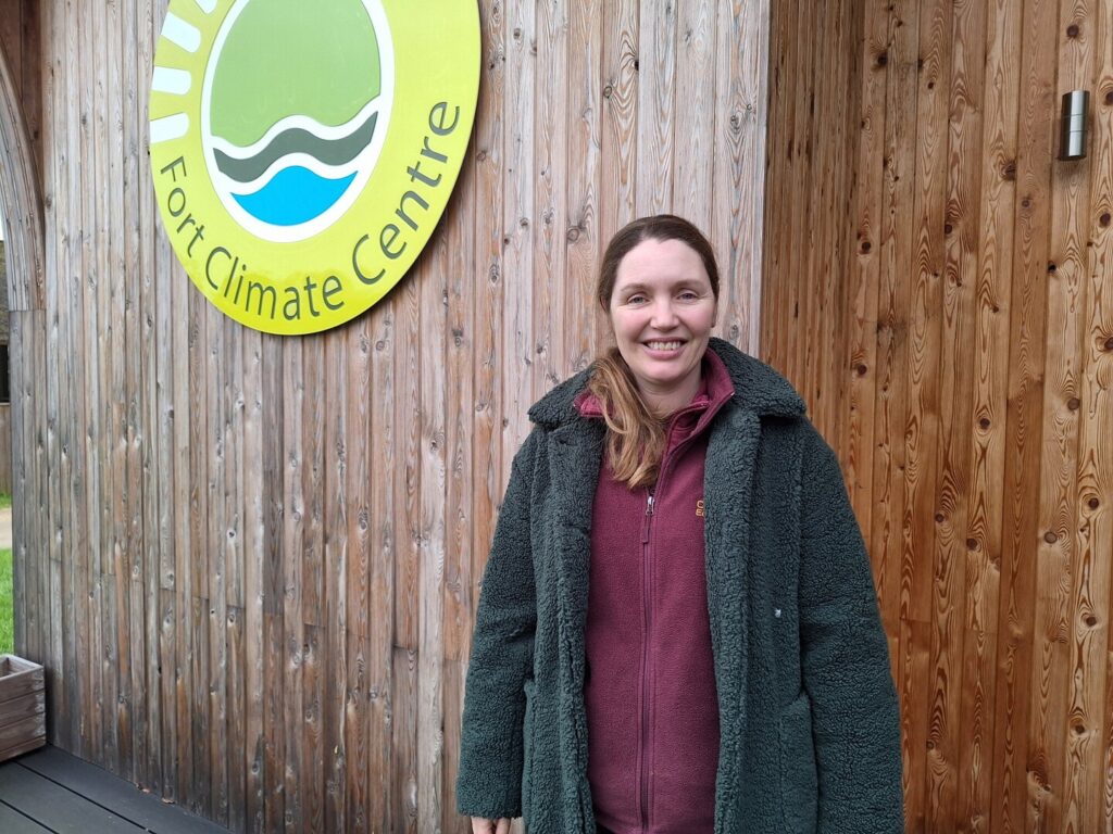A woman stood against a wooden building with a large logo which reads Fort Climate Centre