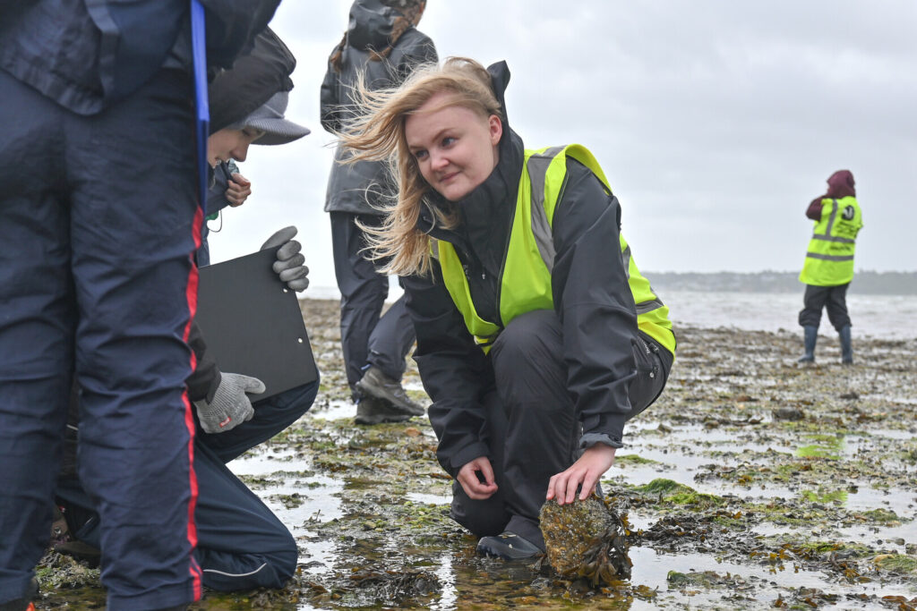 A woman in high-vis kneeling on a beach, lifting a seaweed-covered rock. A young person next to her is looking at the rock. The tide is out and the sky is overcast.