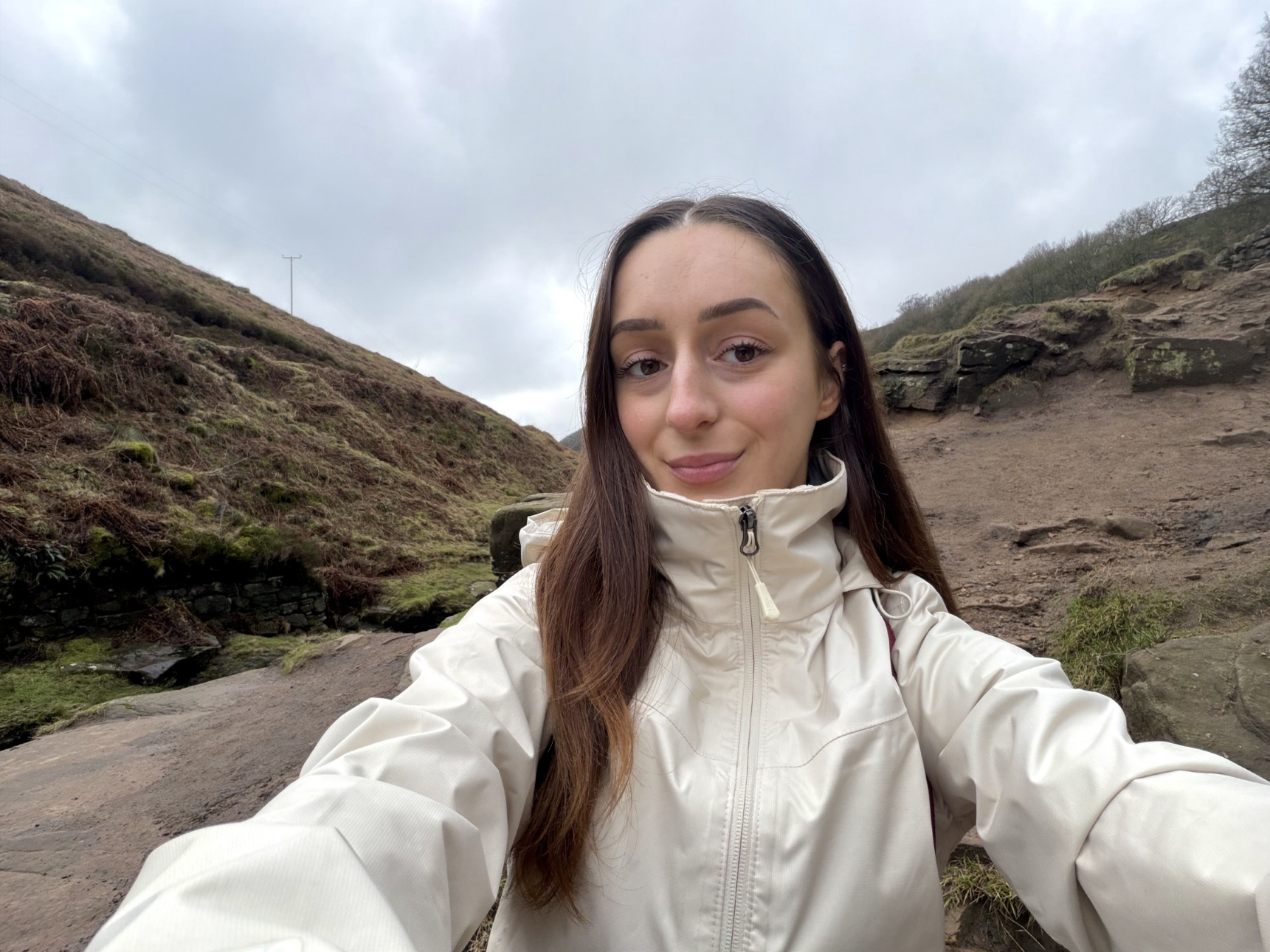 Young woman with brown hair and a white coat taking a selfie with hills in the background.