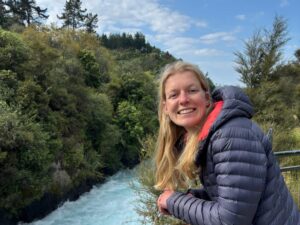 A young woman stands smiling while resting on a railing. Behind her is a forest and a river.
