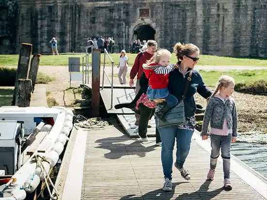 Woman carrying a toddler and holding a young girl's hand while walking off a ferry ramp by a stone fort