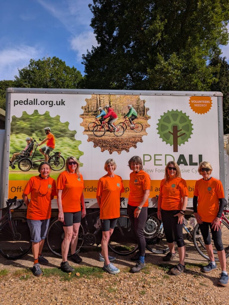 PEDALL - New Forest inclusive cycling's New Forest Loop riders stand in orange PEDALL tops in front of a PEDALL van with trees in the background.