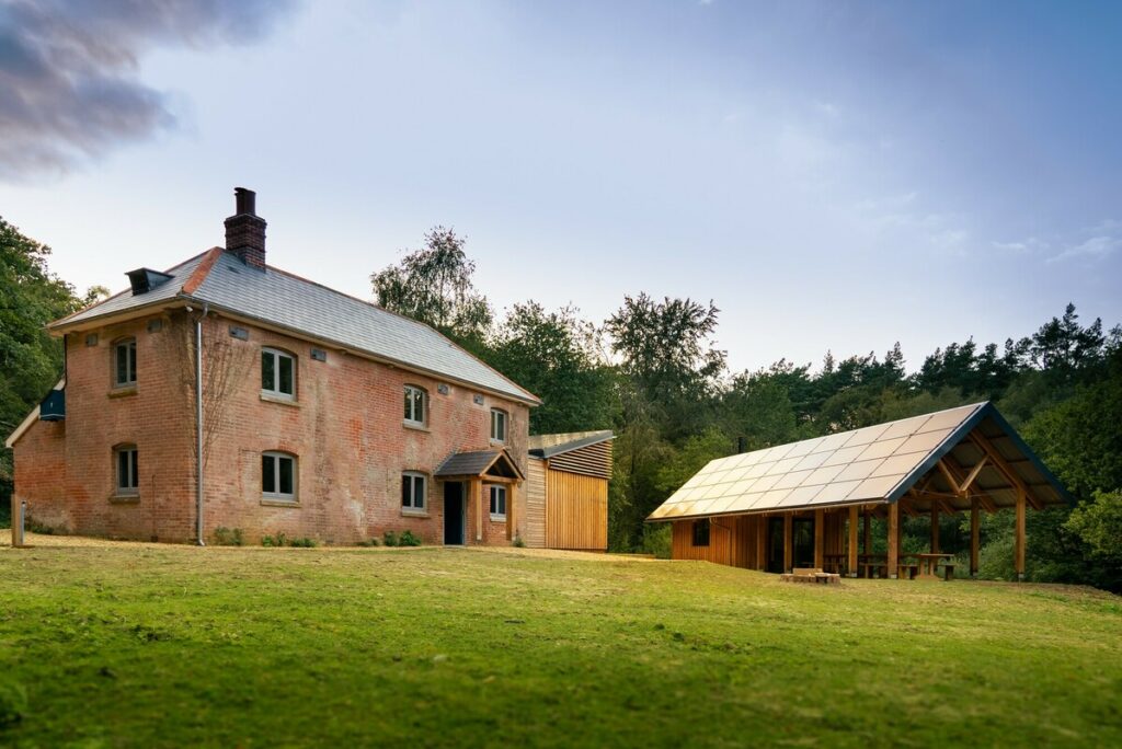 Brick house with a gray metal roof beside a timber pavilion, set on a grassy lawn with trees in the background