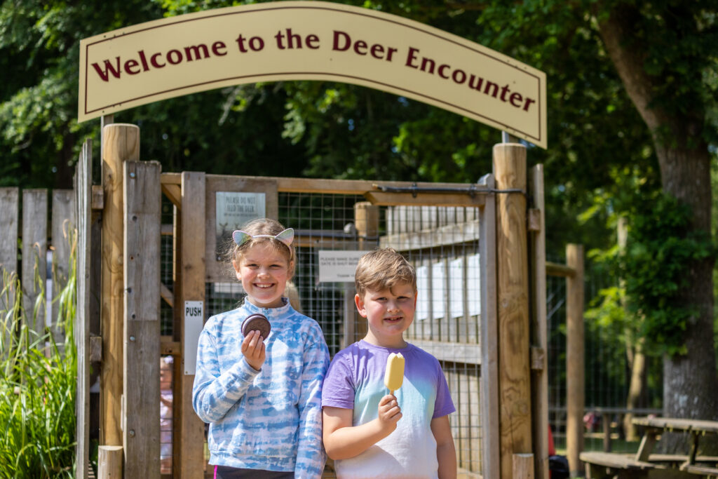 Two children holding ice creams under a Welcome to the Deer Encounter sign at New Forest Wildlife Park entrance in summer