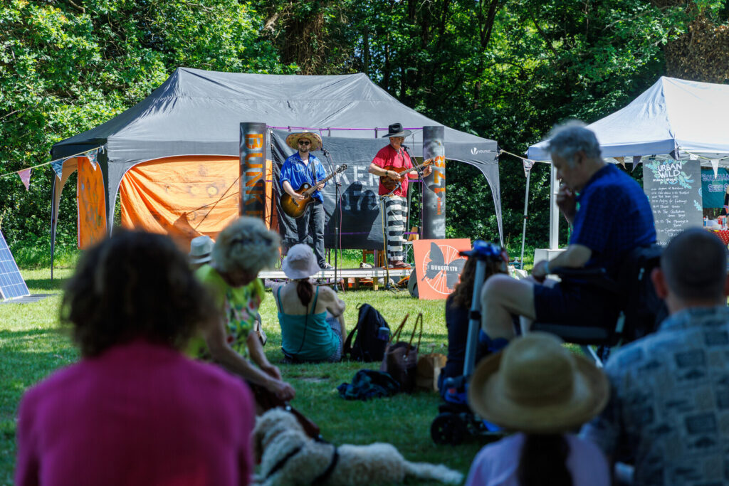 Two singers with guitars on a stage at an outdoor community festival. People are sat in the shade watching them, It's a sunny day.